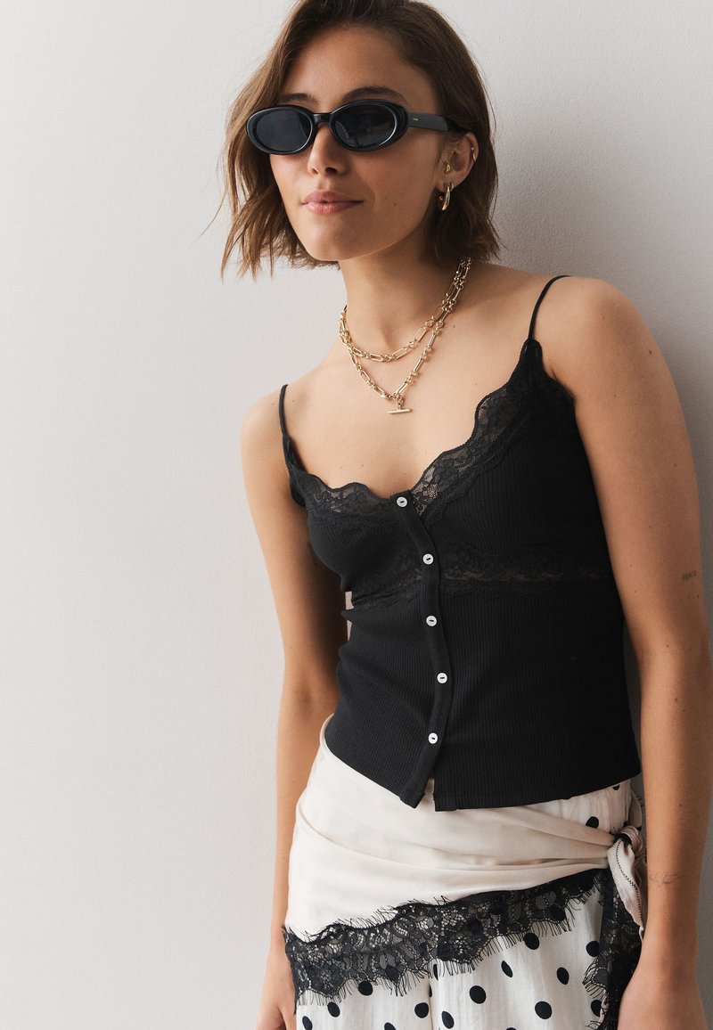 Woman wearing black sleeveless top with buttons, layered gold necklaces, small oval sunglasses, and a white skirt with black polka dots and lace trim.
