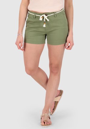 Femme portant un short vert olive avec une ceinture en corde blanche et des sandales beiges, debout jambes croisées contre un fond uni.