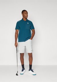 Teal short-sleeve polo shirt with a chest pocket, paired with light gray cargo shorts. White golf shoes with blue accents and dark socks.