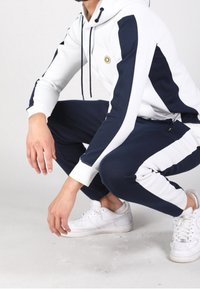 Tuta in bianco e blu navy, con cappuccio, strisce a contrasto, polsini a costine e un logo rotondo sul petto. Abbinato a sneaker bianche.