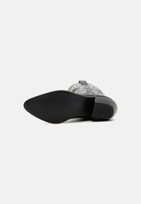 Chaussure à talons noirs avec une semelle lisse et brillante. La tige présente un matériau semi-transparent à motif, avec un talon épais et une sangle réglable.