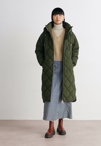 Gequiltte olijfgroene lange jas met een capuchon, gedragen over een beige trui en een spijkerrok. Bruine enkellaarsjes, met minimale hardwaredetails.