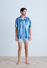 Short-sleeve shirt in blue with abstract wave patterns, made of lightweight fabric. Paired with gray striped shorts. Model standing barefoot.