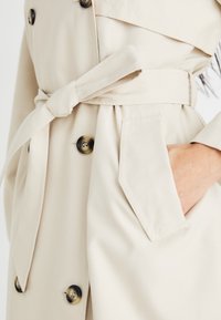 Beige esőkabát övvel a deréknál, dupla gombolással, oldalsó zsebekkel és sima textúrával.