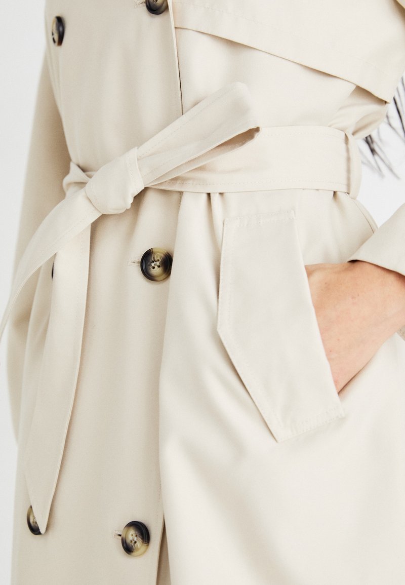 Beżowy płaszcz trench z paskiem w talii, podwójnie zapinany na guziki, z bocznymi kieszeniami i gładką teksturą.