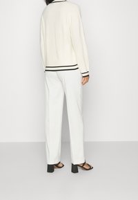 Bílý pletený cardigan s černými pruhy na límci a lemu, v kombinaci s bílými kalhotami a černými sandály na blokovém podpatku.