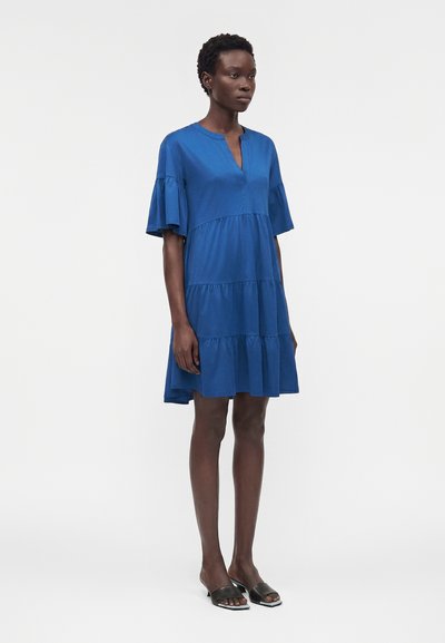 Γυναίκα που φοράει φαρδιά μπλε φόρεμα μήκους μέχρι το γόνατο με στρωματοποιημένες βολάν και κοντά, φουσκωτά μανίκια, συνδυασμένο με μαύρα ανοιχτά πέδιλα slide.