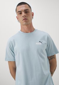 Camiseta de algodón azul claro con cuello redondo y mangas cortas, con un logo negro de Puma en el lado izquierdo del pecho. Textura suave.