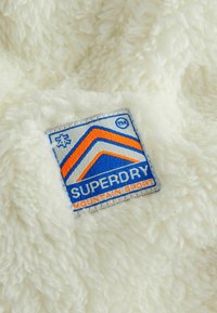 Baltas, pūkuotas audinys su tekstūruotu paviršiumi. Yra mėlyna, oranžinė ir balta siuvinėta etikete, ant kurios užrašyta "SUPERDRY MOUNTAIN SPORT."