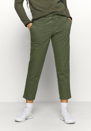 Persoon draagt olijfgroene cropped broek, bijpassende top met lange mouwen, witte sokken en witte sneakers met handen in de zakken.