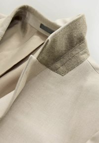 Beige jas met een gestructureerde grijze vilten kraag, met een getailleerd ontwerp en strakke lijnen. De stof lijkt glad en gestructureerd.