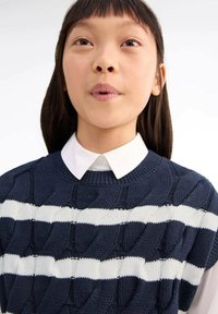 Pleten pulover z mornarsko modrimi in belimi črtami ter teksturno zasnovo, okroglim izrezom, položen preko bele srajce s ovratnikom.