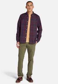 Camicia da uomo a quadri rossa e blu navy con bottoni, in cotone, abbinata a una maglietta color sabbia sotto, pantaloni verde oliva e scarpe marroni.