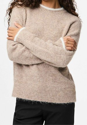 Personne portant un pull beige duveteux avec des bordures blanches aux poignets et à l'encolure, les bras croisés, associé à un pantalon noir.