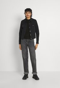 Giubbotto di jeans nero con tasche frontali, indossato sopra un maglione scuro. Abbinato a jeans larghi e sbiaditi arrotolati ai polsi e scarpe da ginnastica nere.