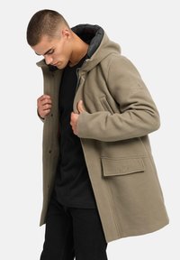 Olivgrön huva jacka gjord av mjukt tyg; har dragkedja, sidofickor och broderad text på ärmen. Svart innerfoder synligt.