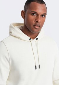 Felpa color crema realizzata in un tessuto morbido. Presenta un cappuccio con cordino e accenti neri, con una vestibilità rilassata. Design semplice e pulito.