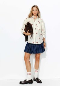 Chemise blanche à boutons avec broderie florale rouge, jupe en denim, sac en franges marron et mocassins marron avec des clous décoratifs. Chaussettes blanches avec texte.