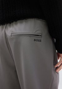 Szürke nadrág sima textúrával, cipzáras hátsó zsebbel és a jobb oldalon jól látható "BOSS" logóval.