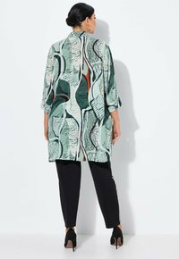Eine Frau, die eine lange, lockere grün gemusterte Jacke, schwarze Hosen mit weißen Streifen und schwarze High Heels trägt, steht vor einer weißen Wand.