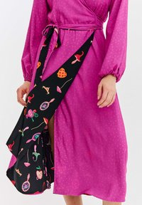 Różowa sukienka kopertowa z długimi rękawami, z czarnym asymetrycznym paskiem ozdobionym kolorowymi wzorami owoców i kwiatów. Sukienka ma teksturę w kropki.