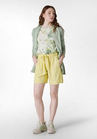Cardigan verde claro sobre una camiseta pálida y estampada con colores suaves. Pantalones cortos amarillos con cintura elástica. Zapatillas beiges con acentos verdes.