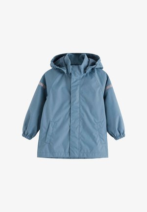 Barn iført blå vandtæt jakke og bukser med sorte knælapper, sorte støvler og en blå strikket hue, stående på hvid baggrund.