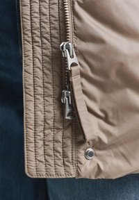 Beige gewatteerde jas met een zilveren rits en drukknoopsluiting, met een gladde textuur en zichtbare stikdetails.