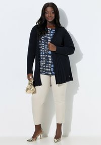 Sort kvinde iført en navy cardigan, mønstret blå top, beige bukser, klare slangeskinds-hæle, holder en lille guldtaske, stående op ad en hvid væg.