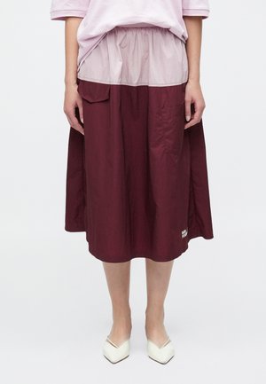 Bordová sukňa strednej dĺžky s nariaseným pásom, s priehľadnou krémovo sfarbenou vrstvou a bočnými vreckami. Textové logo pri leme.