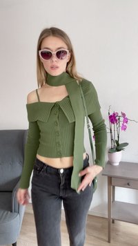 Maglione verde a coste corto con design scollo off-shoulder, chiusura con bottoni e sciarpa attaccata. Indossato con jeans neri, accompagnato da orchidee rosa.