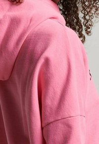 Růžová mikina vyrobená z měkké bavlněné směsi, s uvolněným střihem, oversized kapucí a viditelným šitím na rukávech.