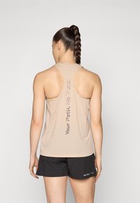 Beige idrettstopp med teksturert stoff og racerback-design, med trykt tekst på baksiden. Kombinert med svarte shorts.