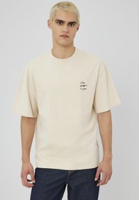 T-shirt beige oversize en coton, avec des manches courtes et un petit motif noir sur la poitrine gauche. Texture simple et lisse.