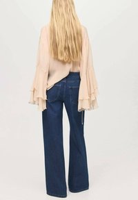 Blouse beige fluide avec des manches cloche superposées, associée à un jean en denim bleu ample, doté de poches arrière et d'une coupe droite.