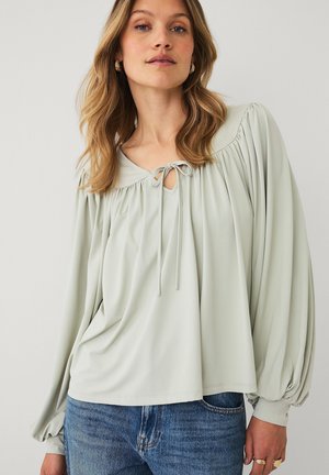 Kvinde iført en sart grøn bluse med samlet halsudskæring og lange puffede ærmer, kombineret med blå denimjeans.