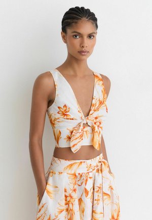 Femme aux cheveux tressés portant un crop top blanc et une jupe assortie avec un imprimé floral orange, debout contre un mur blanc les mains dans les poches.