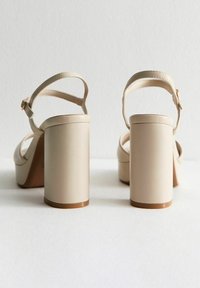 Beige klacksandaler med en slät faux läderfinish, med en bred blockklack och ett tunt ankelband med en metallspänne.