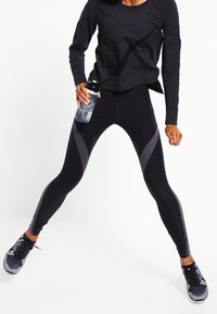 Leggings atléticos negros con acentos grises, hechos de un material elástico. Combinados con una camiseta de manga larga oscura y zapatillas negras. Botella de agua en mano.
