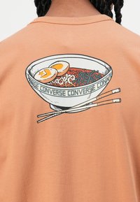 Persiku krāsas T-krekls ar grafiku, kurā attēlota nūdeles ar olām un ēdamstickām, detalizēta ar melnajām kontūrām un tekstu.