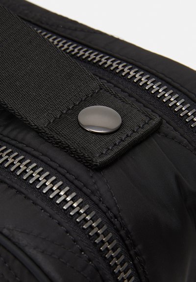 Detaliu al unei genți negre din material textil, evidențiind un fermoar argintiu, un buton magnetic negru și o curea texturată, reflectând un design modern și accente funcționale.