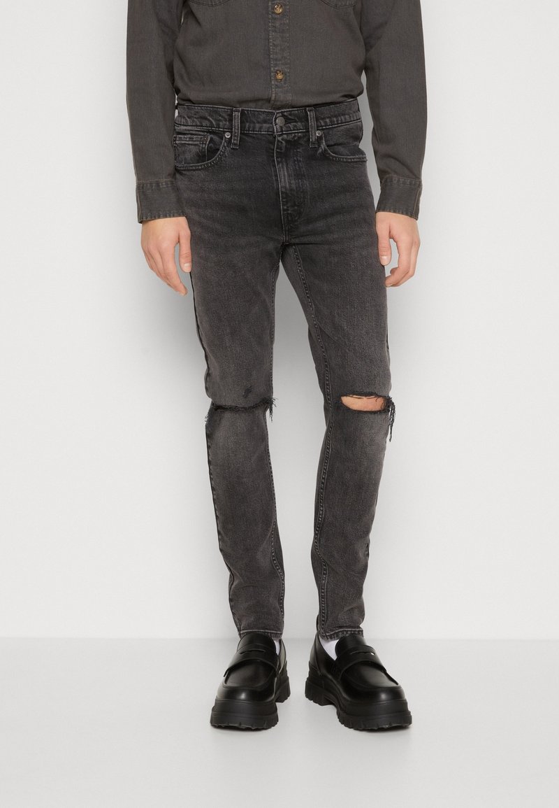Personne portant une chemise foncée à boutons, un jean noir déchiré avec des trous aux genoux et des chaussures noires, debout devant un fond uni.
