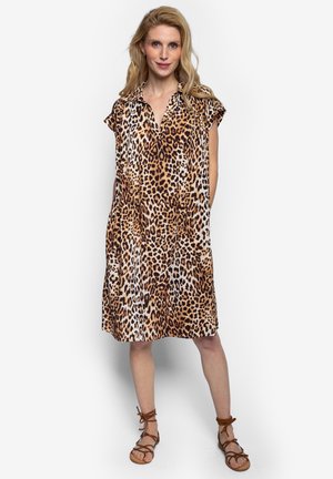 Blondynka stojąca w sukience w panterkę do kolan z kołnierzykiem i krótkimi rękawami, nosząca brązowe sandały na paski, na białym tle.