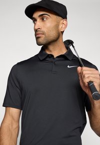 Polo shirt de golf Nike noir avec un col, manches courtes et un logo blanc. La personne tient un club de golf, portant une casquette noire.