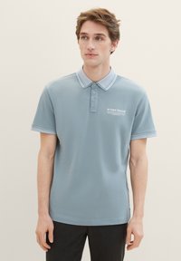 Svetlo modra polo majica z ovratnikom, tremi gumbi in rebrastimi detajli. Ima majhen logotip in besedilo na prsih.