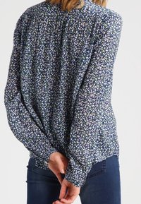 Blumenbluse mit blauem Hintergrund, die mit kleinen, bunten Blumen verziert ist. Lange Ärmel, geraffte Rückendetails und lockere Passform.