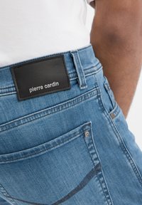 Jean en denim bleu clair avec une patch en cuir noir portant "pierre cardin" en argent. Comprend des poches arrière standard et des détails de couture subtils.