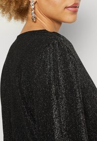Μαύρη μπλούζα με υφή, λαμπερό ύφασμα και φουσκωτά μανίκια. Διαθέτει στρογγυλή λαιμόκοψη. Ορατό ασημένιο εντυπωσιακό σκουλαρίκι στη μοντέλο.