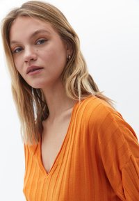 Top in maglina a coste arancione con scollatura a V profonda e tessuto testurizzato. Caratterizzato da maniche corte e una silhouette aderente. Sfondo bianco uniforme.