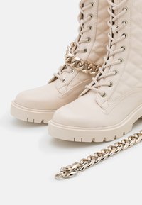 Stivali beige con lacci e design trapuntato, dotati di suola spessa e un accento di catena argento removibile. Texture in pelle liscia.